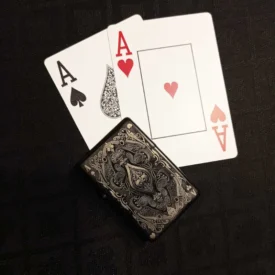 Schwarzes Feuerzeug mit detailliertem Spielkarten-Design (Pik-Ass) und zwei Pokerkarten (Pik-Ass und Herz-Ass) auf schwarzem Tisch.